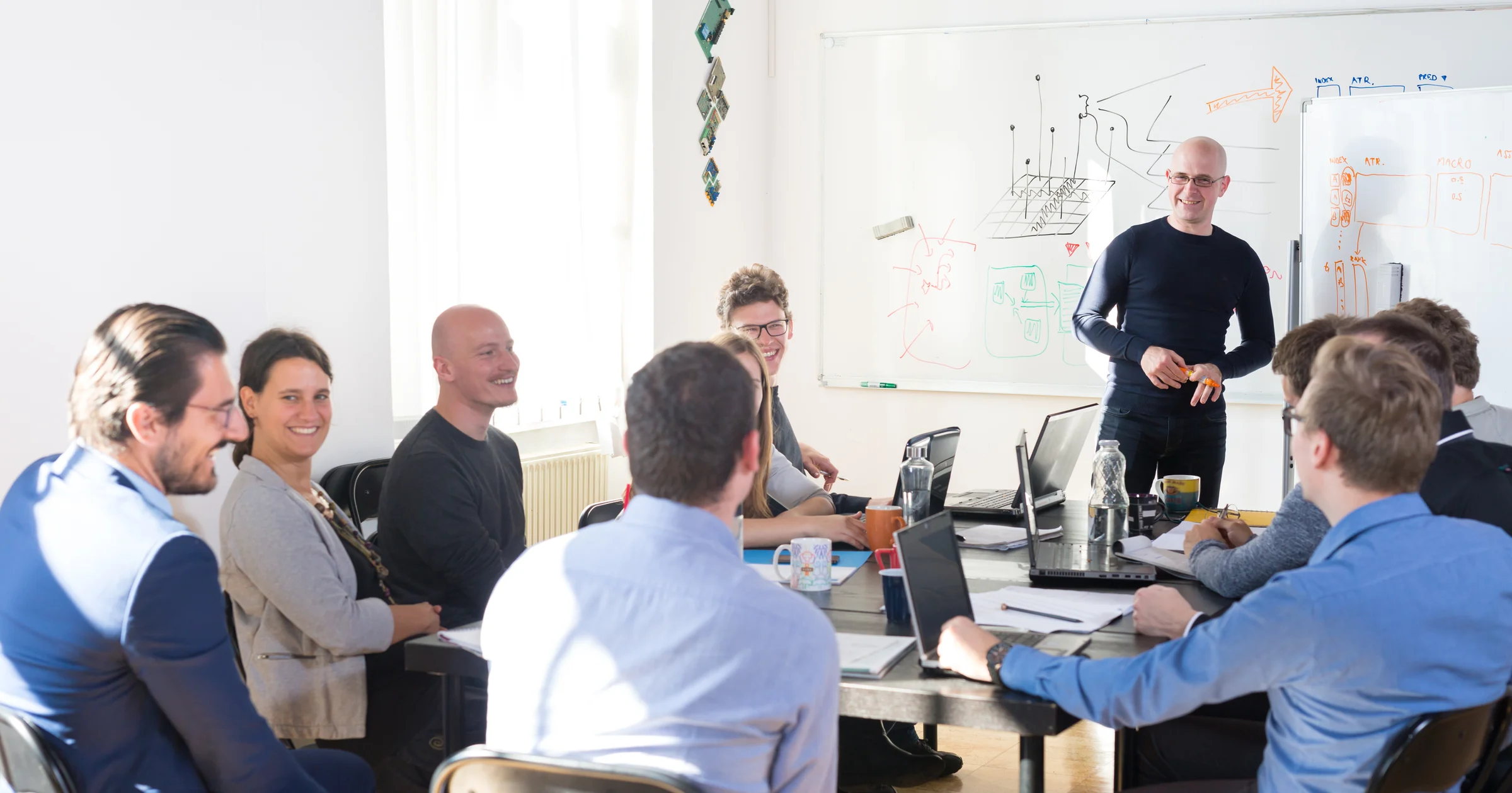 IT team meeting around laptops, with technical diagrams on a whiteboard